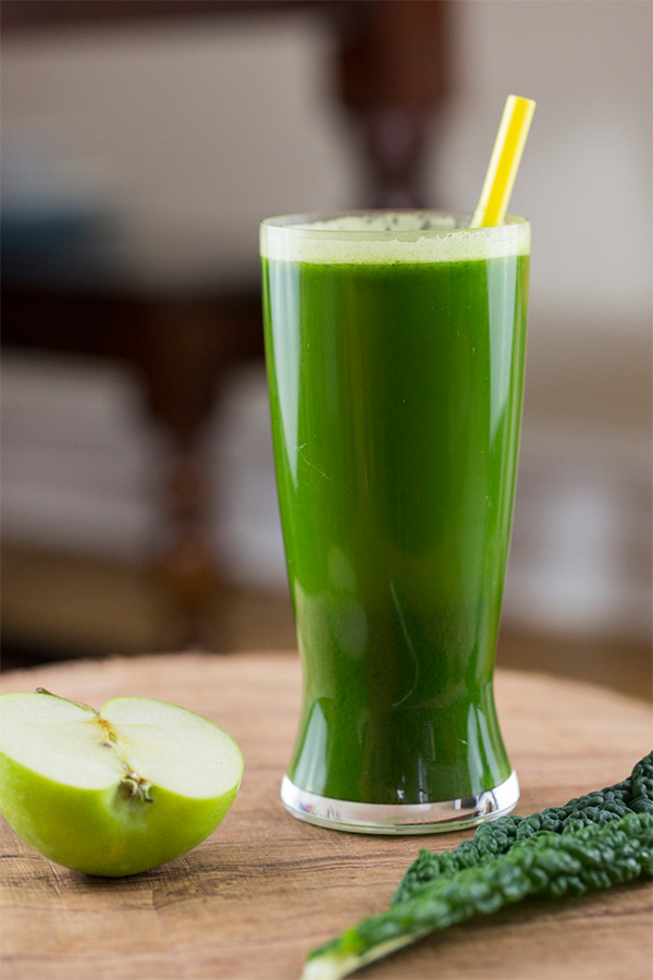 A photograph of a green smoothie in a glass with a green apple and kale